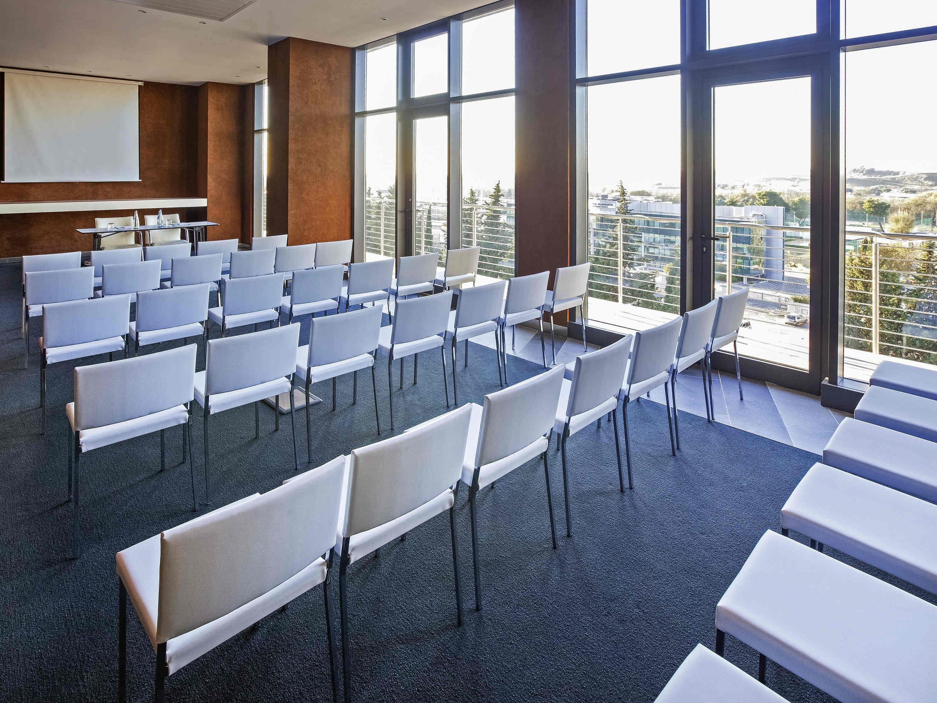 A conference room with rows of chairs, a projection screen, and large windows offering a city view.