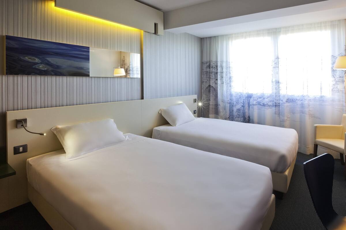 Quarto de hotel com duas camas, um quadro na parede, uma janela com cortinas e uma mesa com uma cadeira.