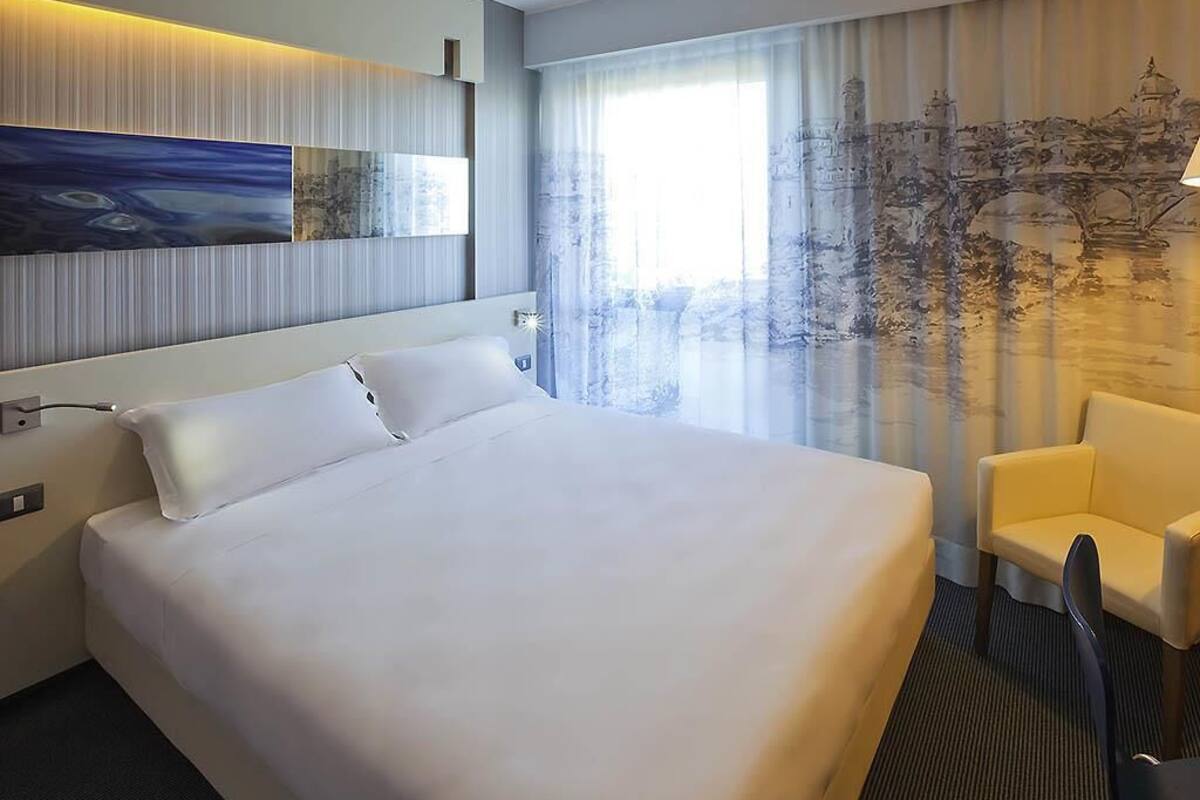 A hotel room with a large bed, a chair, a painting on the wall, and a window with a view.