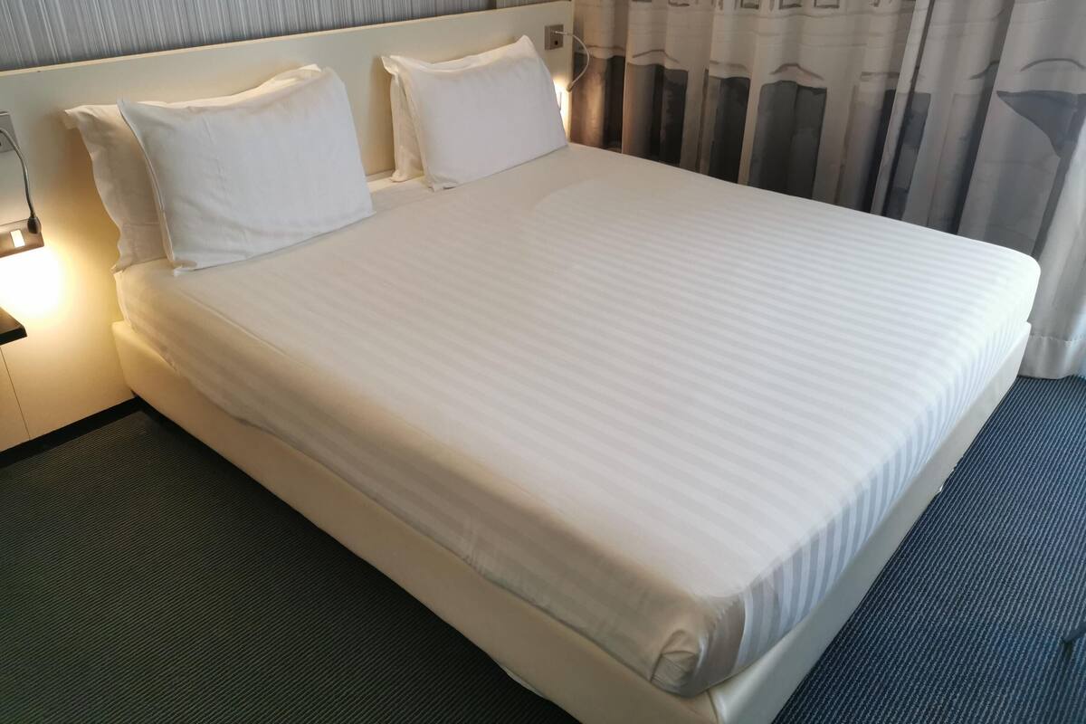 Een hotelkamer met een groot bed, twee kussens, een nachtkastje met een lamp en een raam met gordijnen.