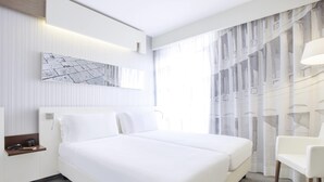 Une chambre d’hôtel moderne avec un grand lit, une table de chevet avec une lampe, une chaise et une vue sur la ville à travers de fines rideaux.