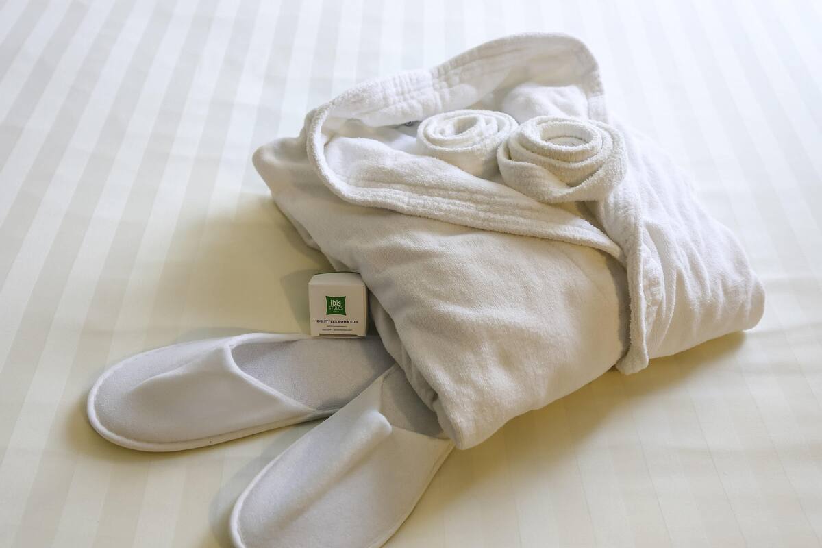 Een netjes gevouwen witte handdoek, twee witte slippers en een klein wit doosje met de tekst 'IBIS STYLES ROMA EUR' op een gestreept dekbed.