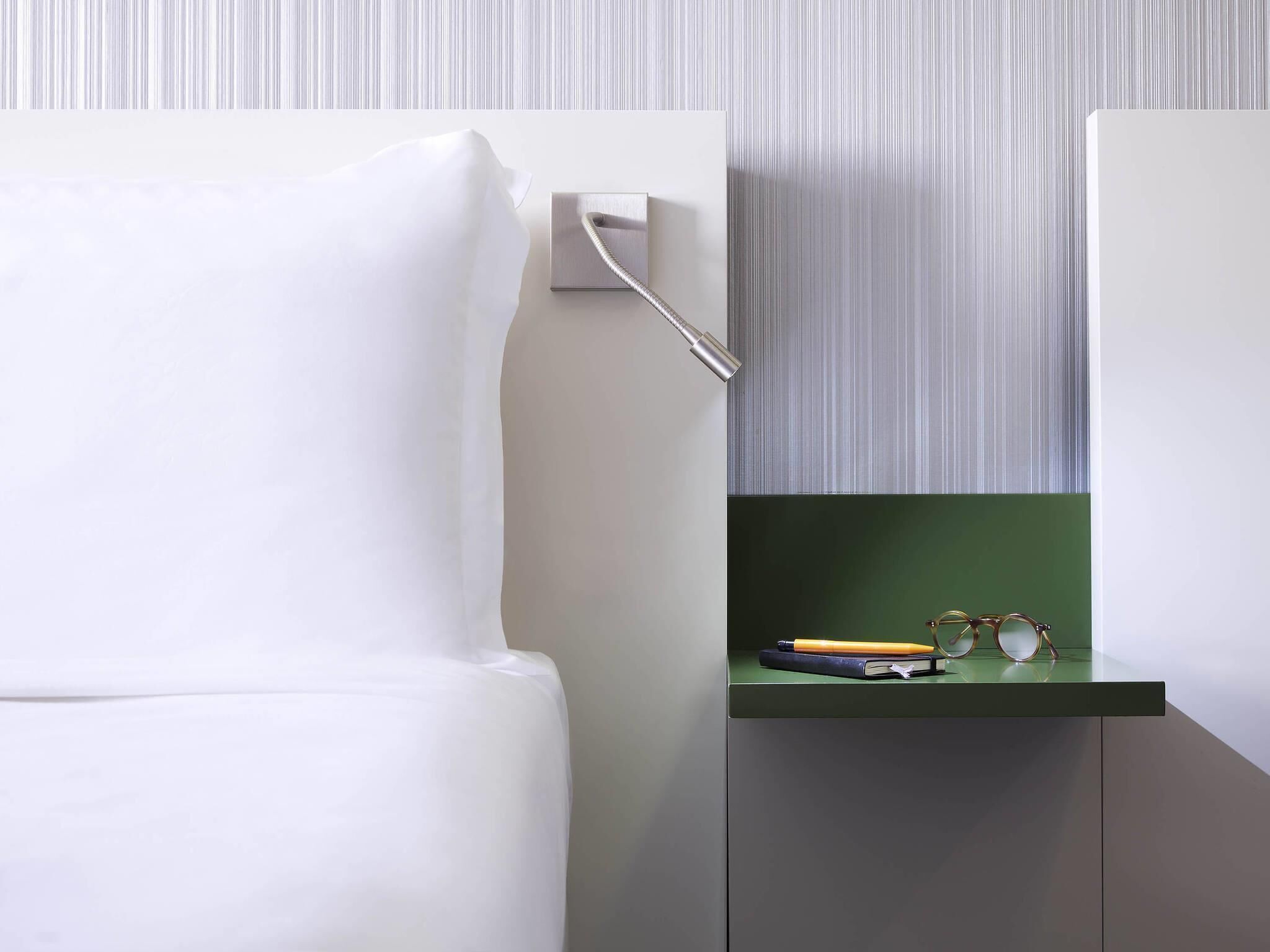 A modern room with a white bed, a green nightstand with glasses and a book, and a striped wallpaper background.