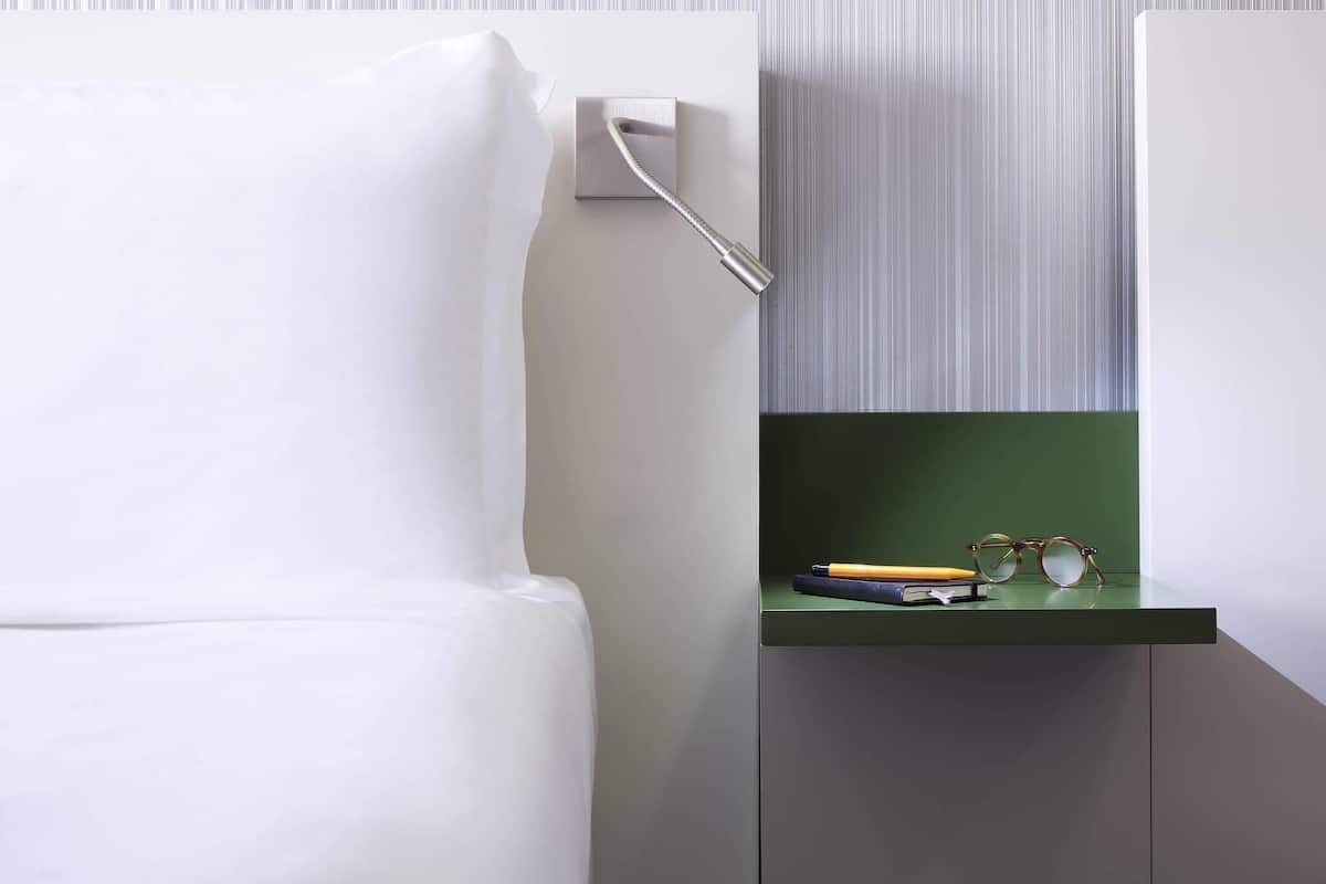 Une chambre moderne avec un lit blanc, une table de chevet verte sur laquelle se trouvent des lunettes et un livre, et un papier peint à rayures en arrière-plan.