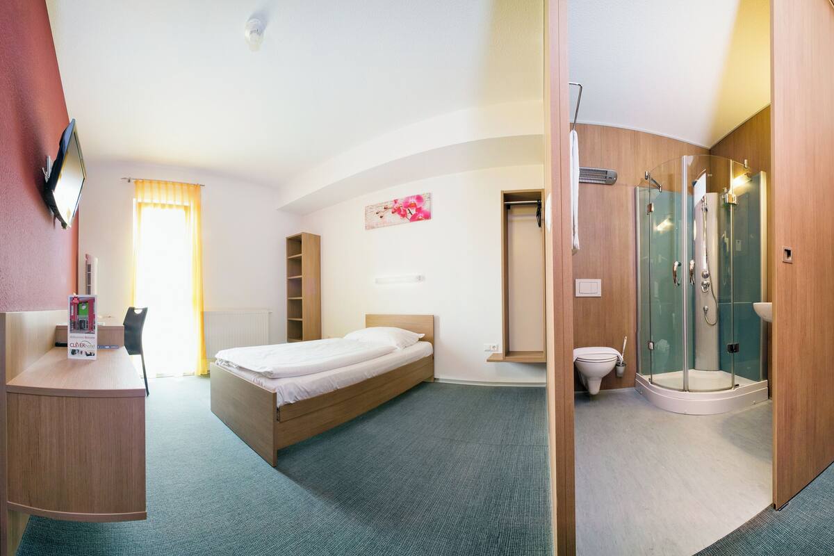 A hotel room with a bed, a desk, a shower, and a toilet.
