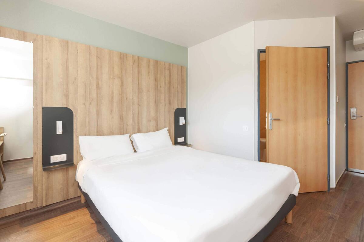 A modern hotel room with a wooden headboard, a large bed, a bedside table, and a wardrobe.