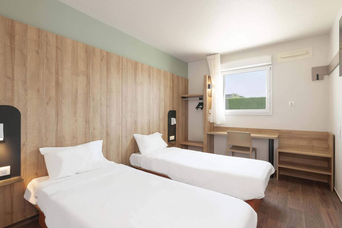 A room with two single beds, a desk, a chair, and a window with a view of a green field.