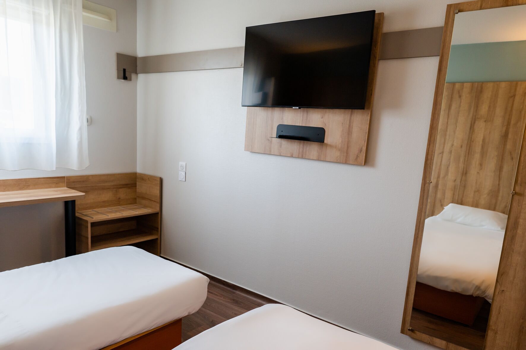 Ein Hotelzimmer mit an der Wand befestigtem Fernseher, einem Holztisch, einem Bett und einem Kleiderschrank.