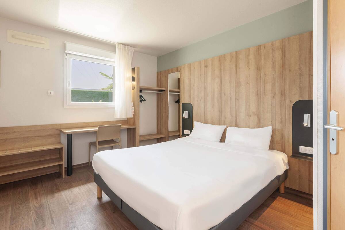 A hotel room with a large bed, a desk, a chair, a wardrobe, and a window.