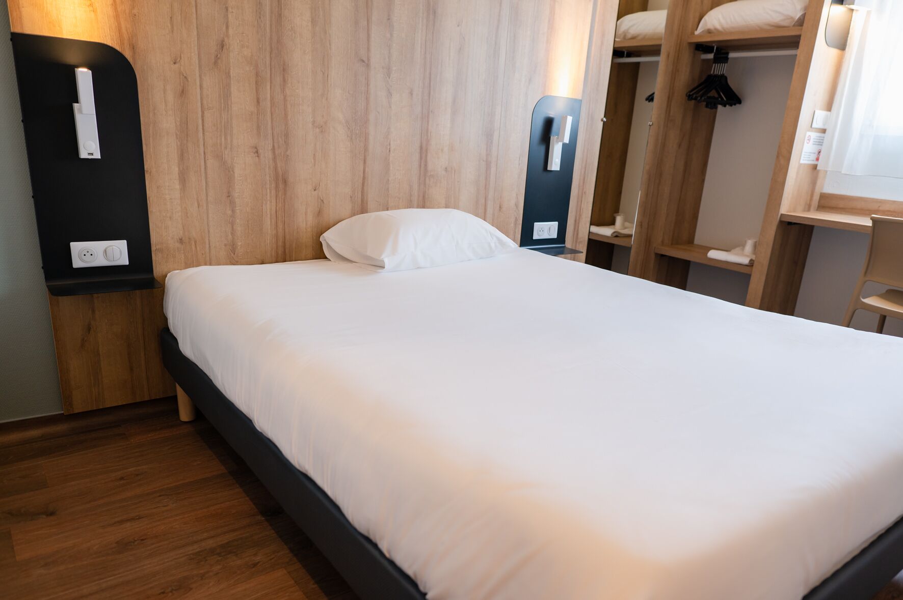 A neatly made bed with a white mattress and pillow, a wooden headboard, and a black bedside table with a light switch.