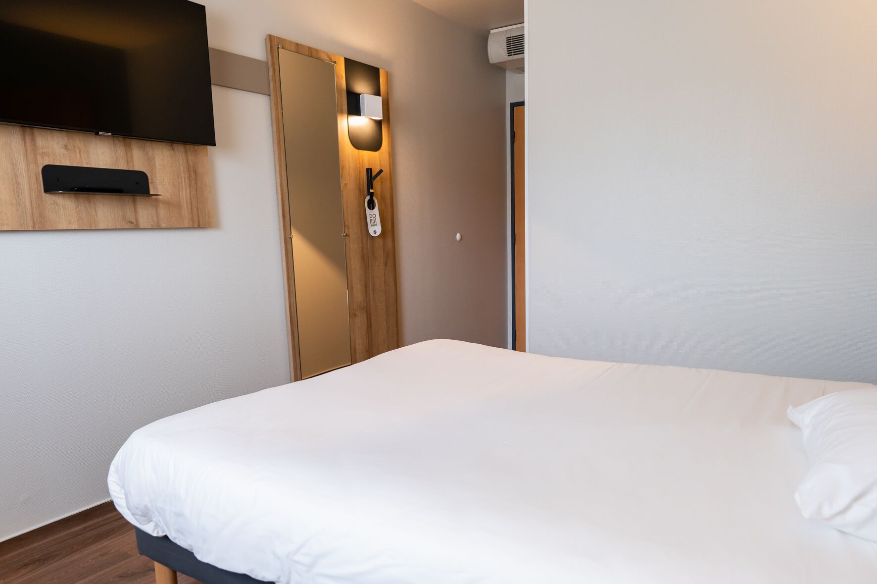 Habitación de hotel con una cama, televisor montado en la pared, armario de madera y una puerta con interruptor de luz.