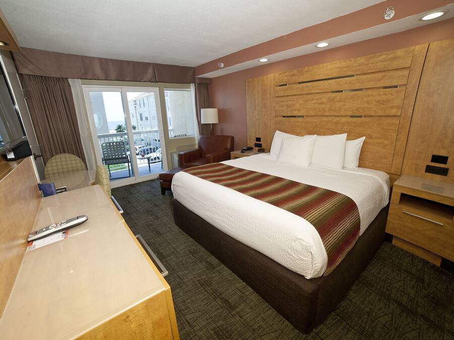 Habitación de hotel con una cama grande, un escritorio con control remoto, una silla, una mesita de noche y vistas al balcón exterior.