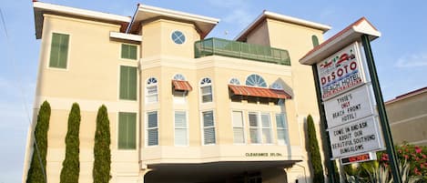 A hotel with a clear entrance, a signboard advertising the Desoto Beach Hotel, and a parking area with several cars.