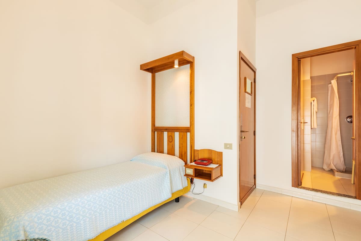 A small bedroom with a single bed, a wooden nightstand, a mirror, and a wooden door leading to a bathroom.