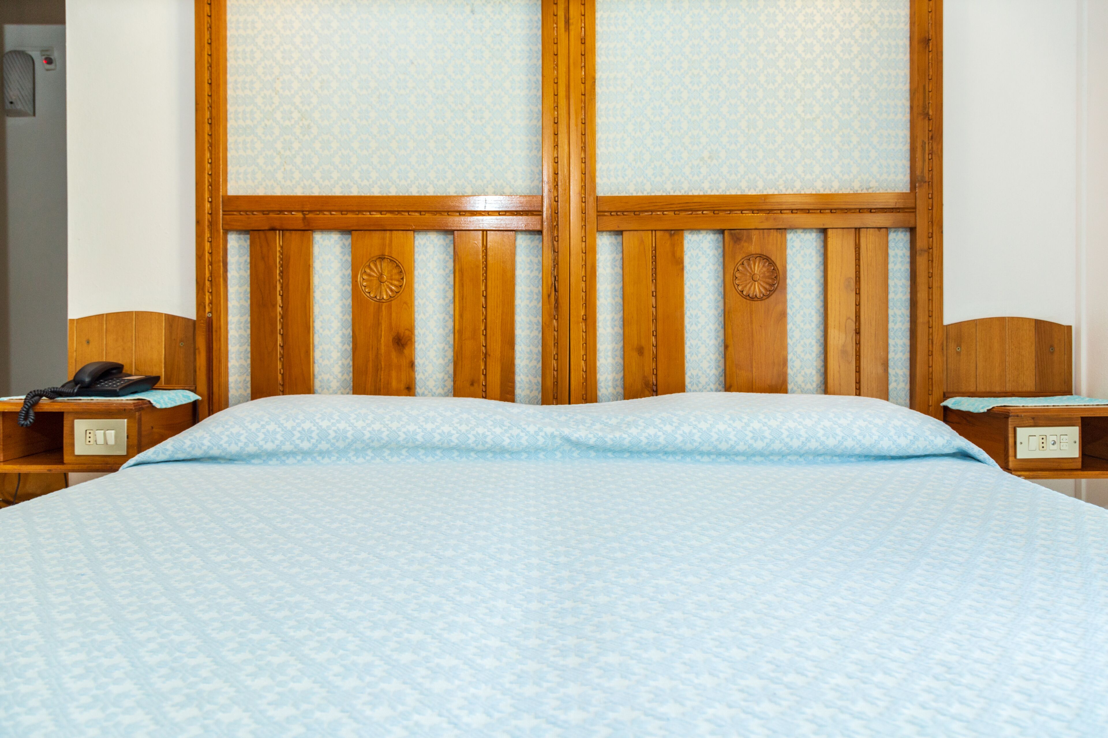 Une chambre d’hôtel avec une tête de lit en bois, un lit recouvert d’une couvre-lit à motifs, un téléphone sur la table de chevet et une petite table de nuit avec une lampe.