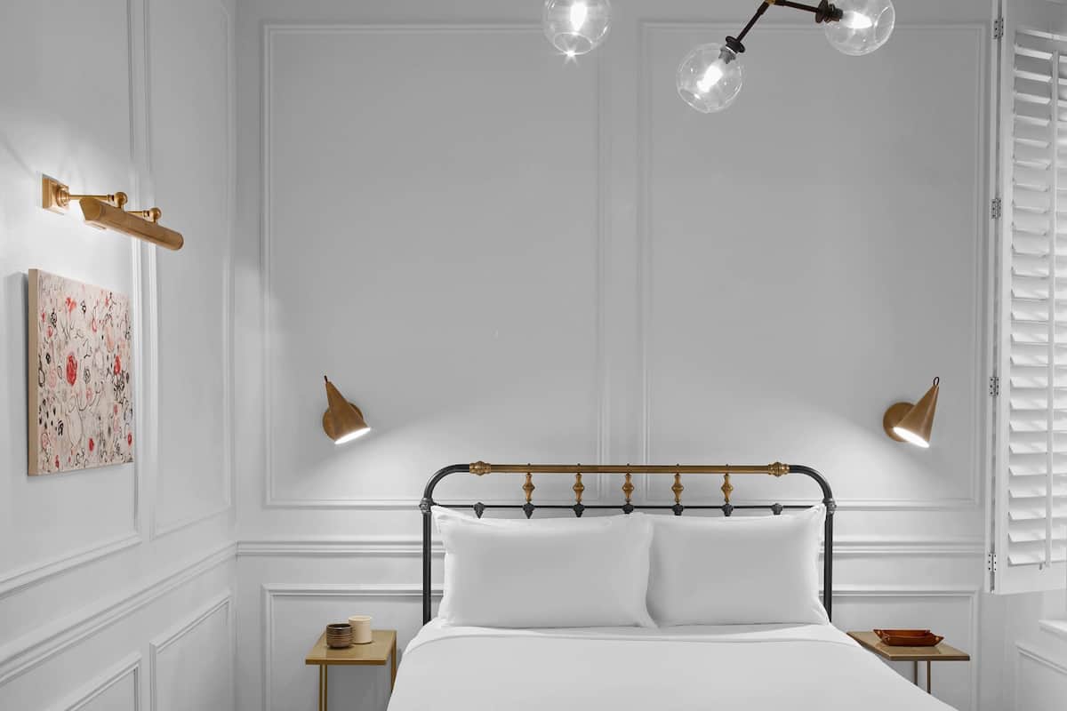 A bedroom with a white bed, a wooden nightstand, a bedside table, a wall-mounted lamp, and a ceiling light fixture.