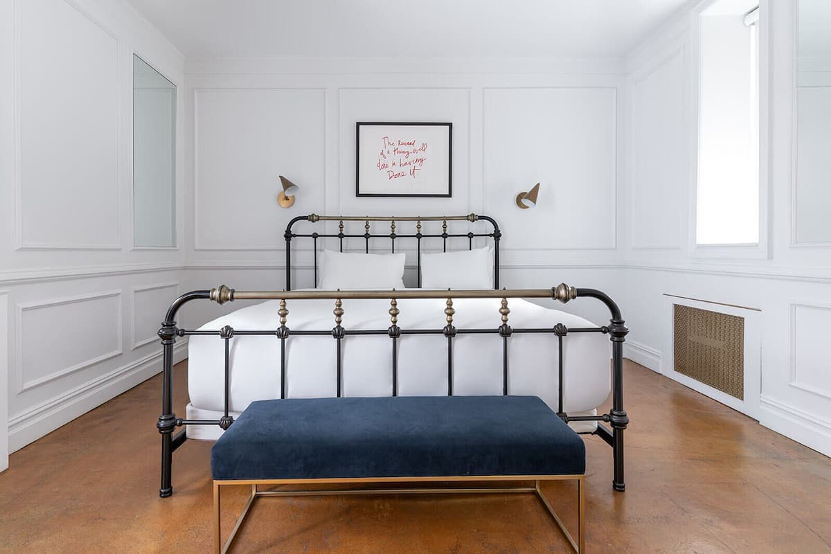 A bedroom with a metal bed frame, white bedding, a blue bench, and a framed quote on the wall.