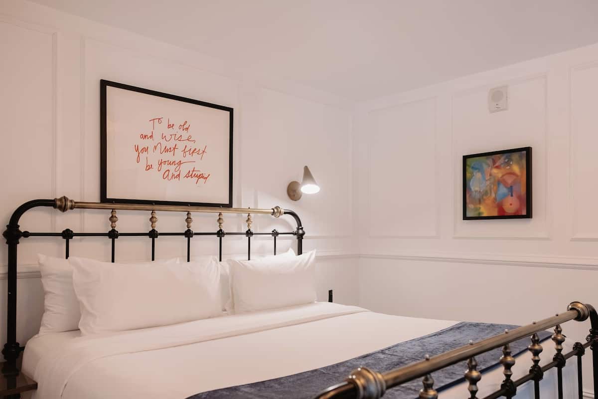 A neatly made bed with a headboard, a wall-mounted light, and two framed artworks.