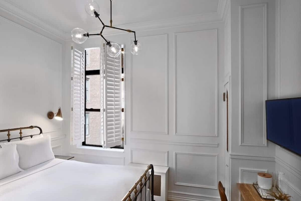 A bedroom with a bed, a window with shutters, a wall-mounted TV, and a bedside table with a plant.