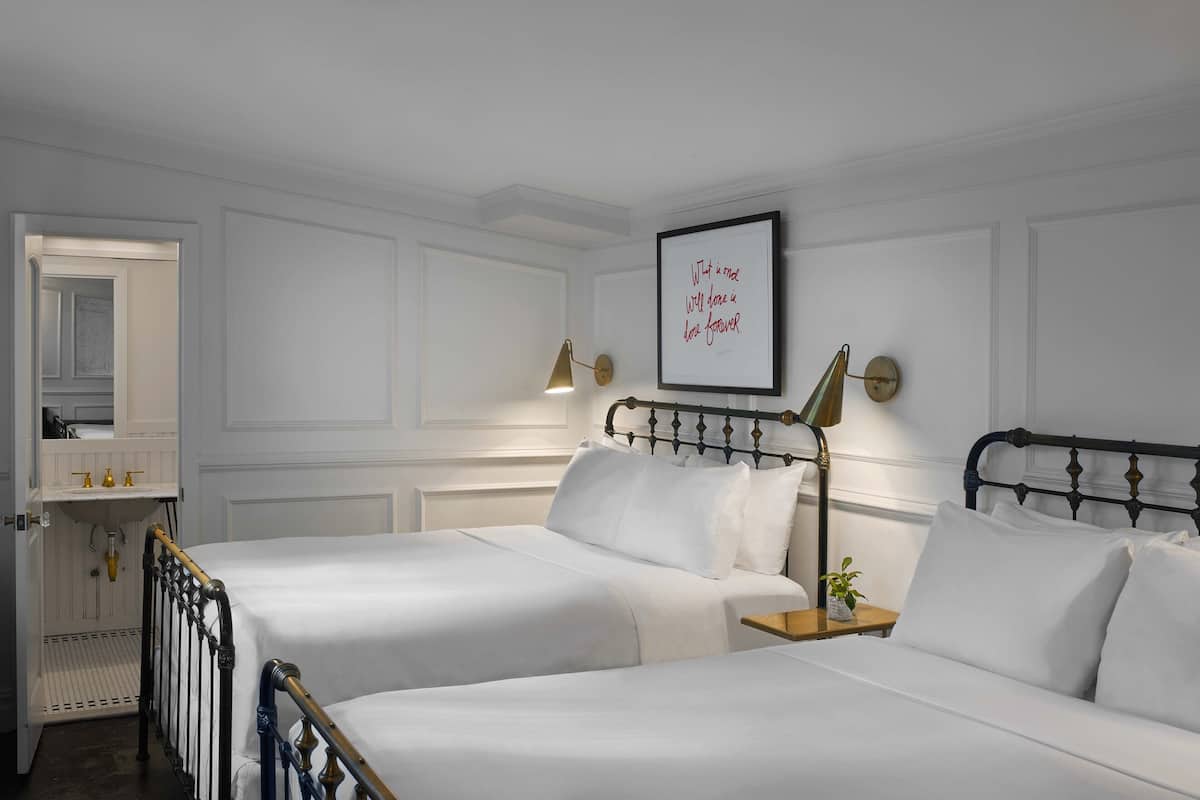 A hotel room with two beds, a bedside table with a lamp, a framed quote on the wall, and a bathroom with a mirror and sink.