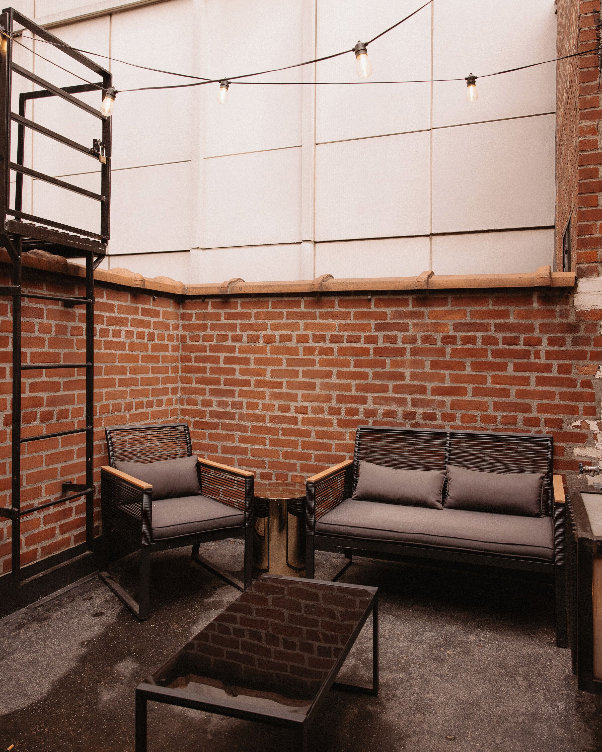 Eine Ziegelwand, zwei Stühle, ein Sofa, ein kleiner Tisch und Lichterketten in einem Innenhof.