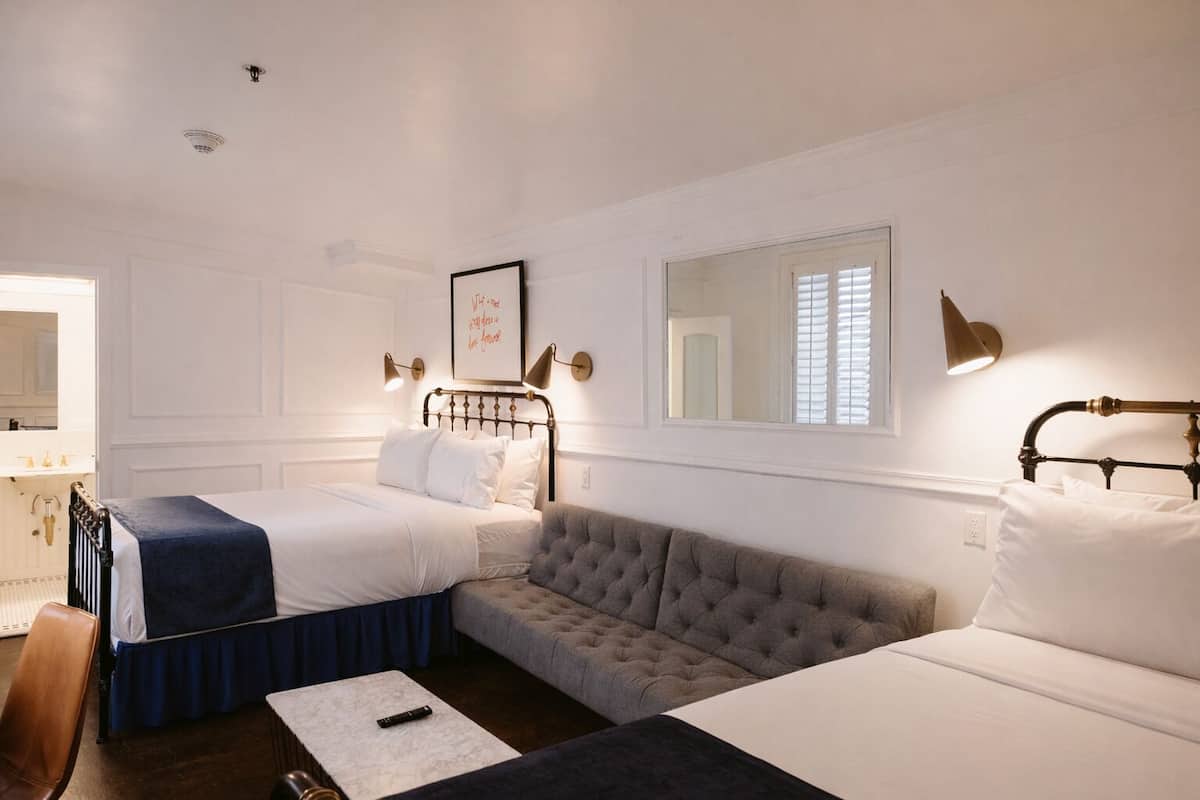A hotel room with two beds, a sofa, a small table, a chair, a mirror, and wall-mounted lamps.