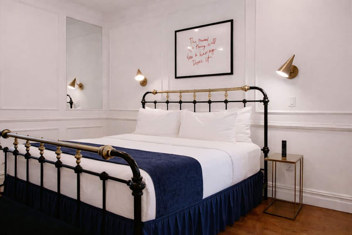 A neatly made bed with a blue skirt, a gold and black metal frame, and white bedding. There is a framed quote on the wall, a small bedside table with a remote, and a wall-mounted light fixture.