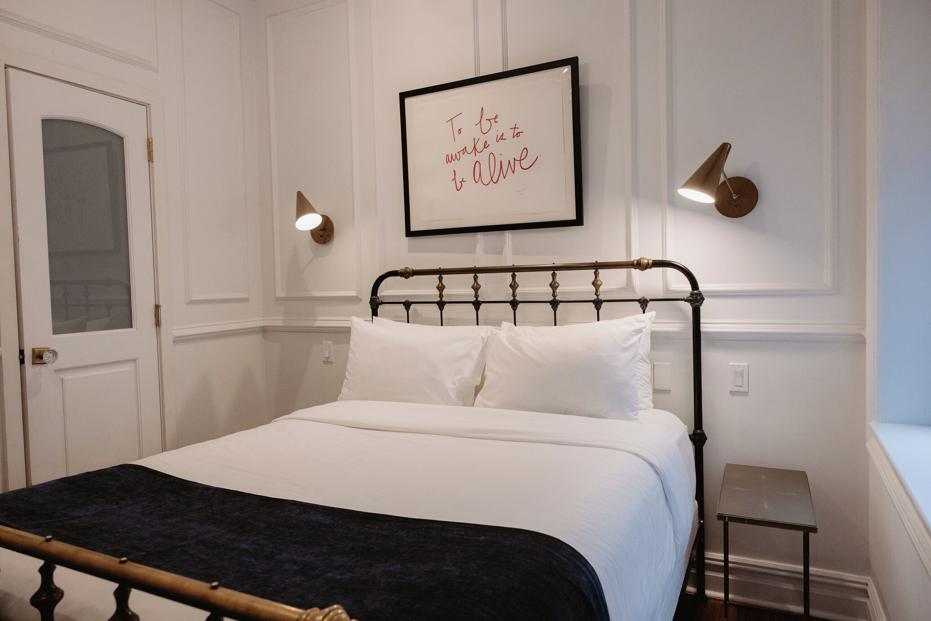 A neatly made bed with a dark throw blanket, a framed inspirational quote on the wall, and two wall-mounted lamps.