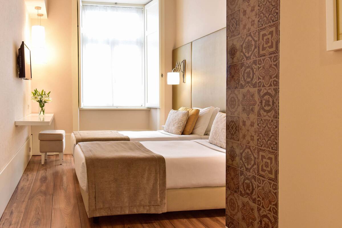 Una camera d'albergo con due letti, un comodino, un tavolino, una finestra e piastrelle decorative alle pareti.