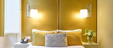 A hotel room with a bed, two bedside tables, a nightstand, a lamp, and a vase with flowers.