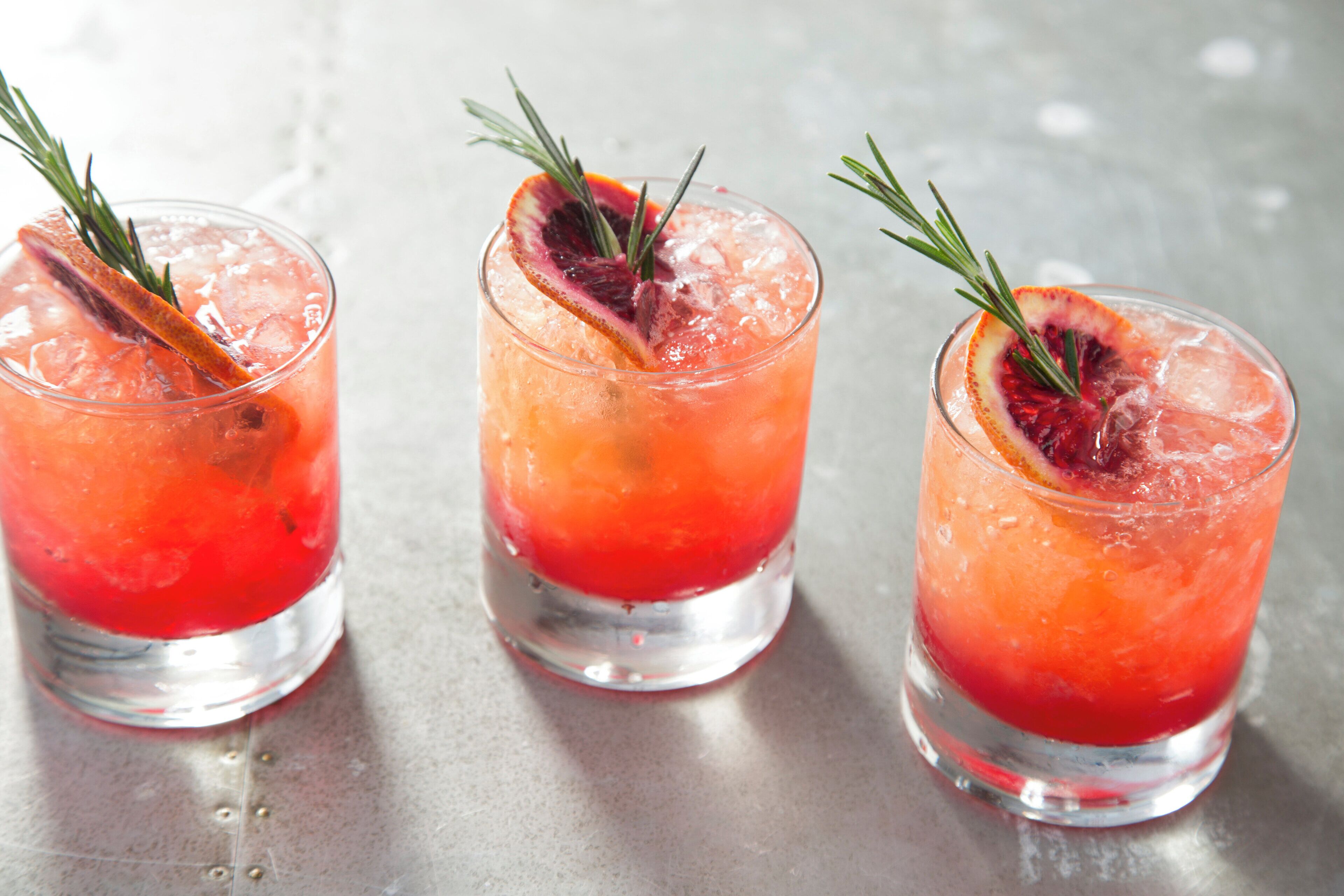 Tres vasos de una bebida roja con hielo y un adorno, sobre una superficie metálica.