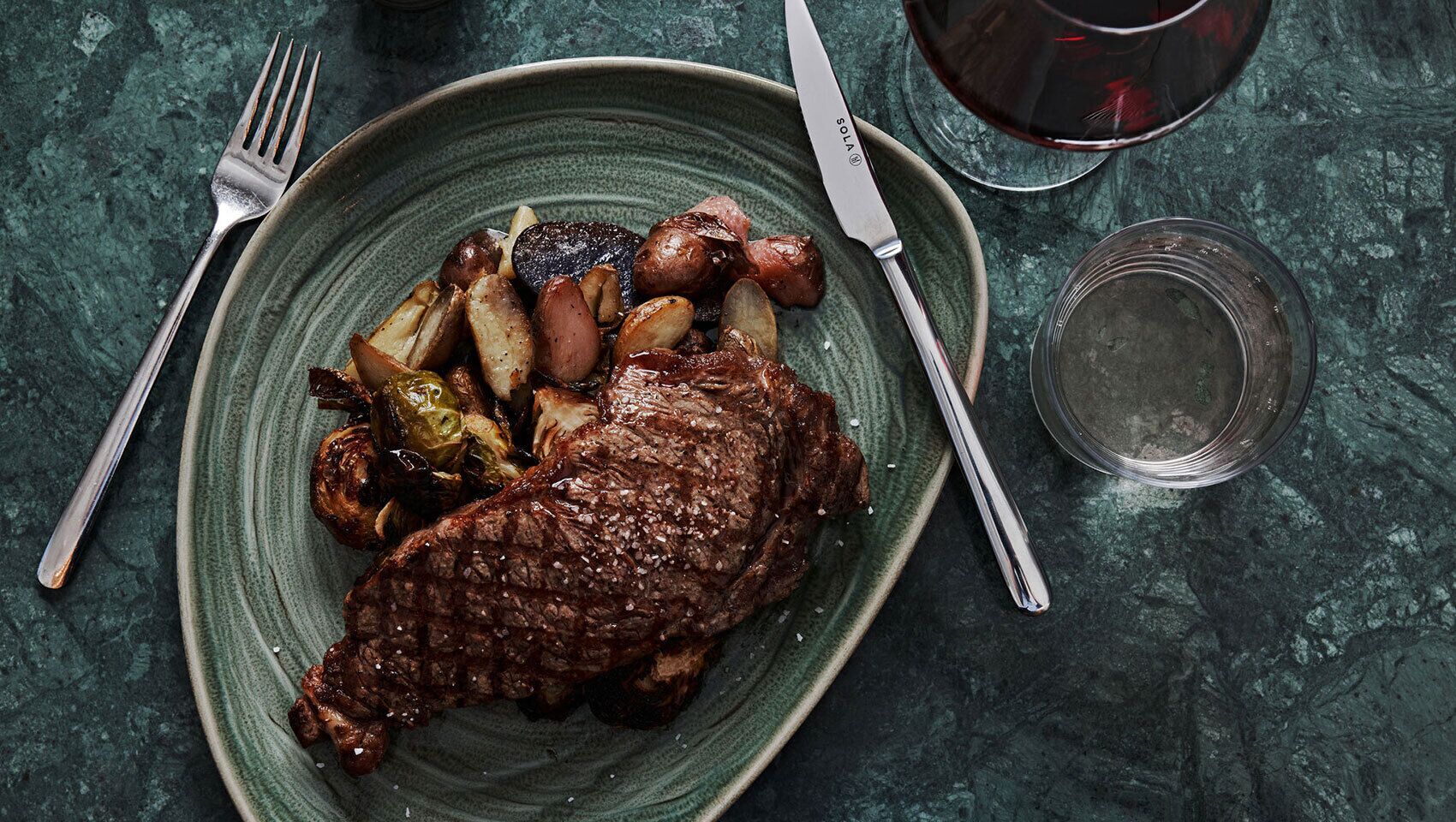 A plate with a steak and roasted vegetables on a textured green plate, accompanied by a fork, knife, and a glass of red wine.