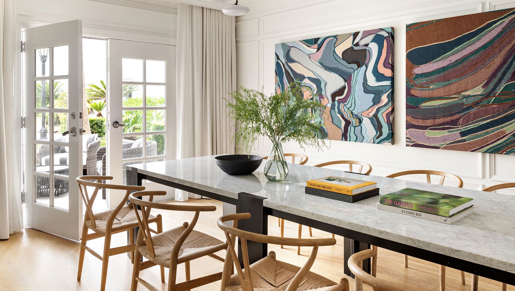 Un comedor con una mesa de mármol, sillas de mimbre, un jarrón con una planta y dos cuadros abstractos en la pared.