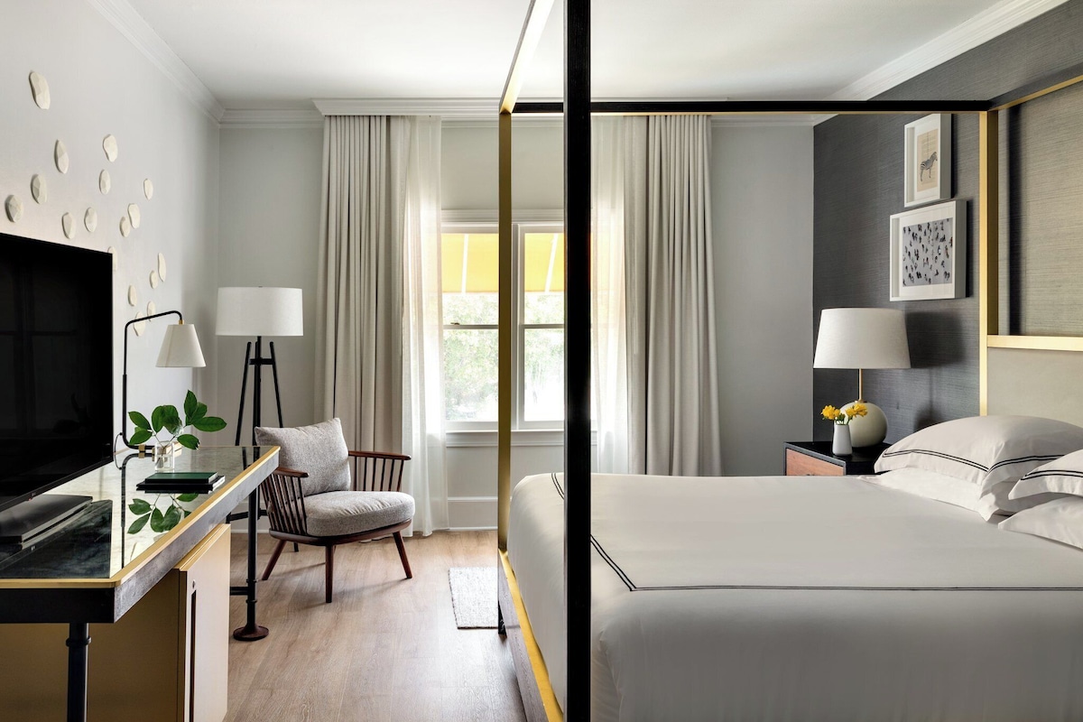 A modern hotel room with a bed, a desk with a TV, a chair, and a window with curtains.