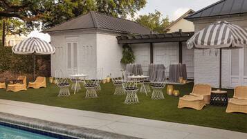 A poolside area with white wicker chairs and tables, striped umbrellas, and a brick building with a pergola.