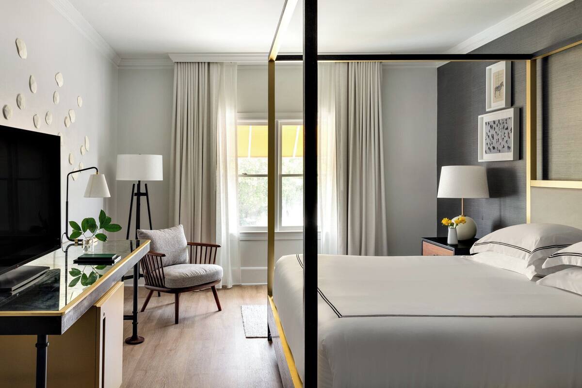 A modern hotel room with a bed, a desk with a TV, a chair, and a window with curtains.
