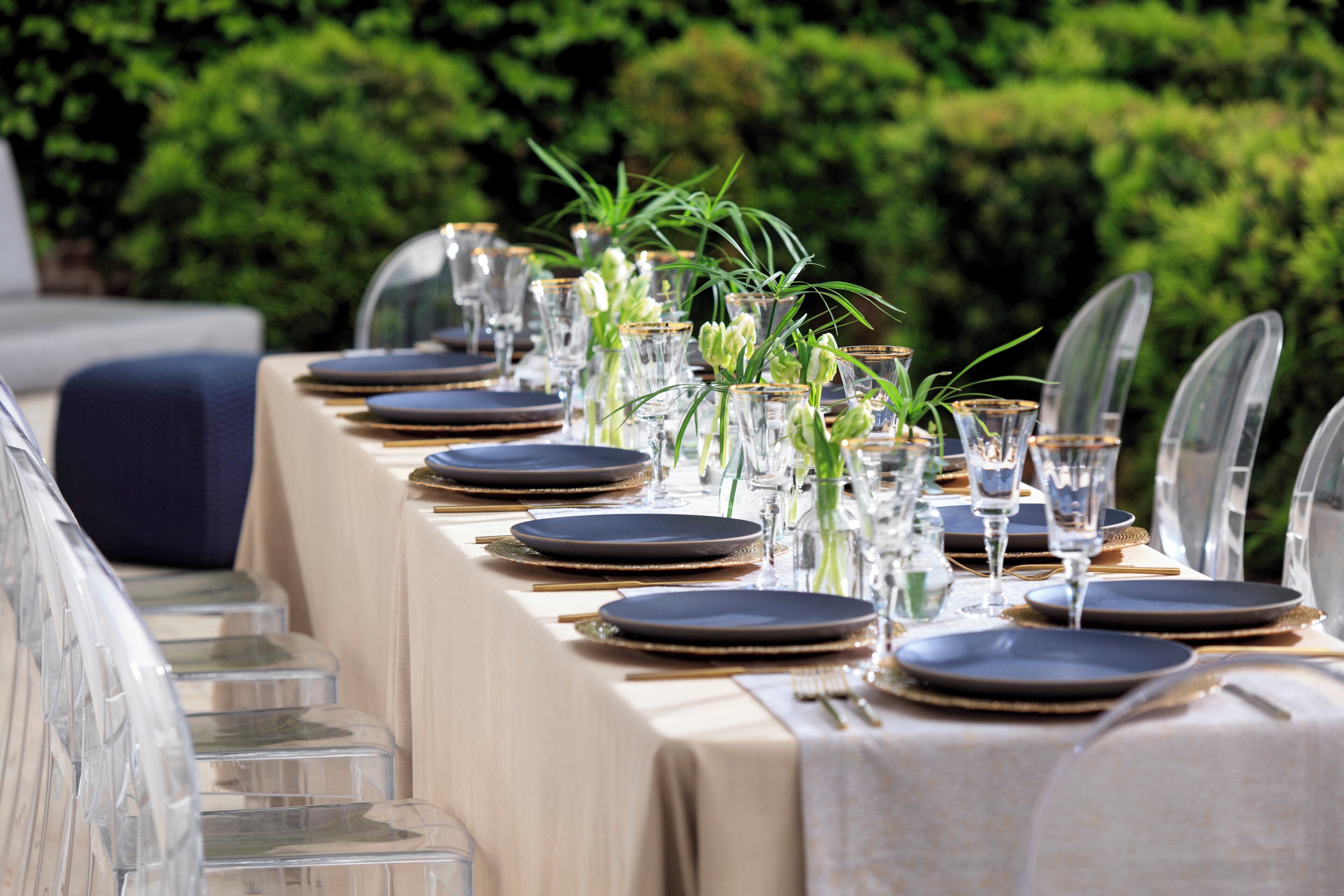 A table set for a formal outdoor dining event with clear glassware and plates, surrounded by greenery.