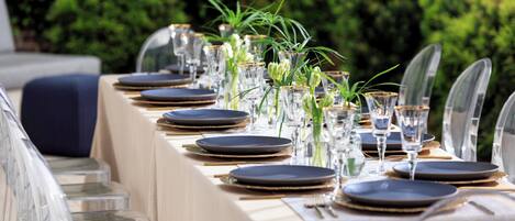 A table set for a formal outdoor dining event with clear glassware and plates, surrounded by greenery.