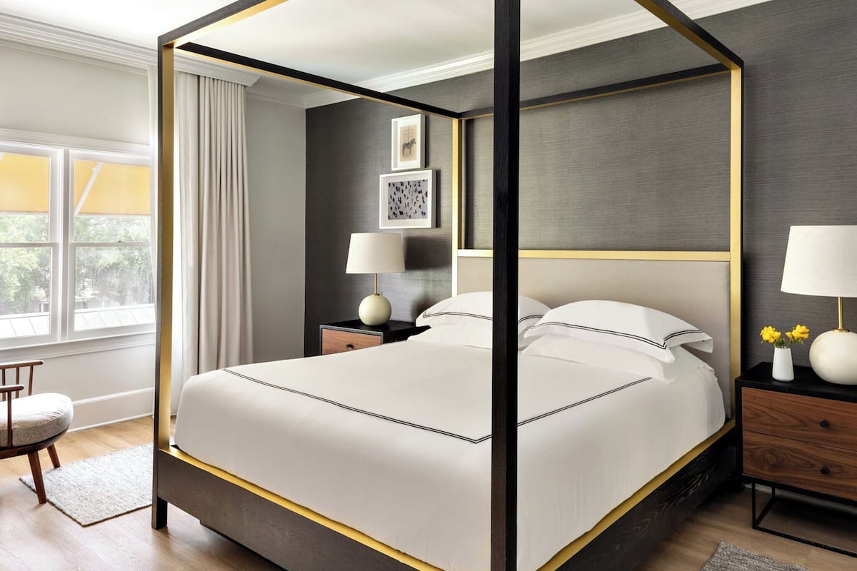 A four-poster bed with a dark frame and white bedding, a nightstand with a lamp, and a window with curtains.
