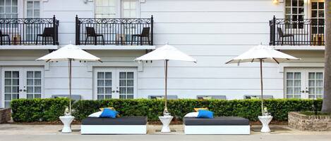 A poolside area with lounge chairs and umbrellas, a hedge, and a building with balconies in the background.