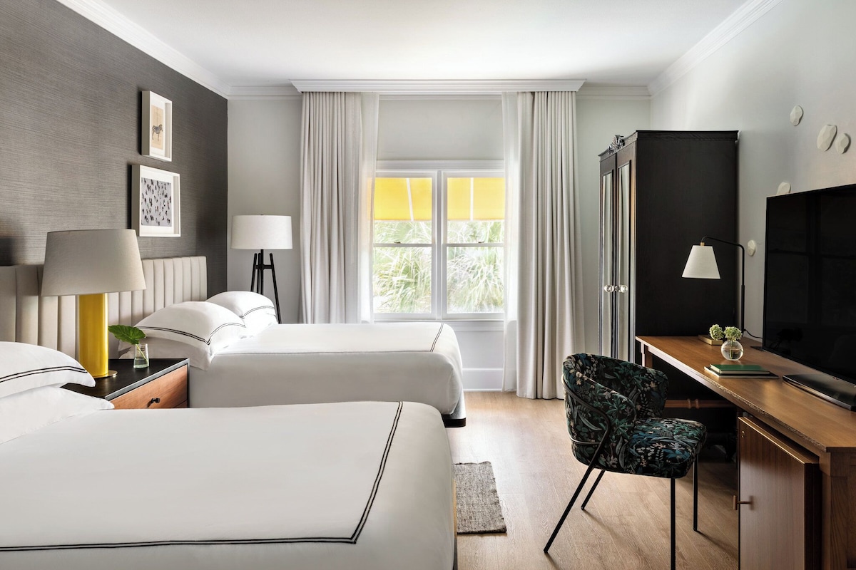 A hotel room with two beds, a desk, a TV, and a window with curtains.