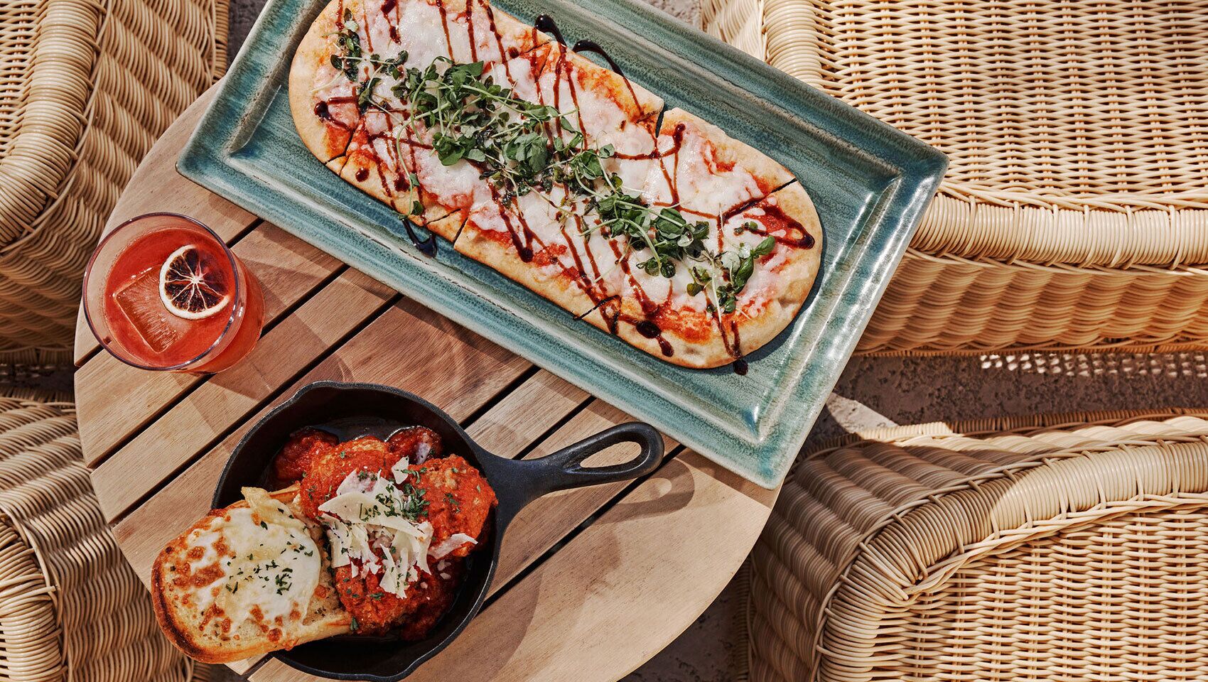 A pizza on a blue plate, a glass of red drink, and a dish of pasta on a wooden board.