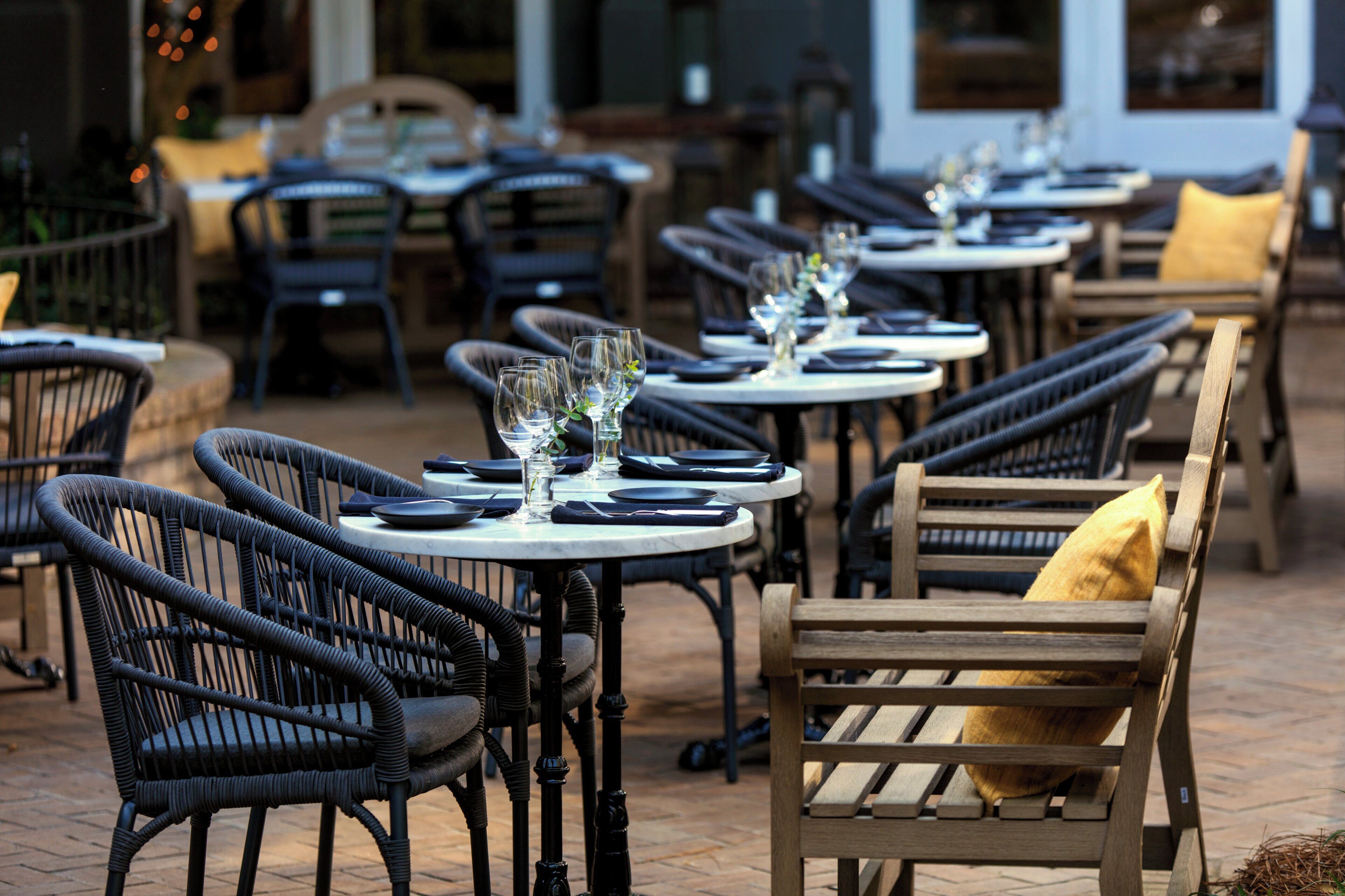 An outdoor dining area with round tables, wicker chairs, and a wooden bench with a cushion.