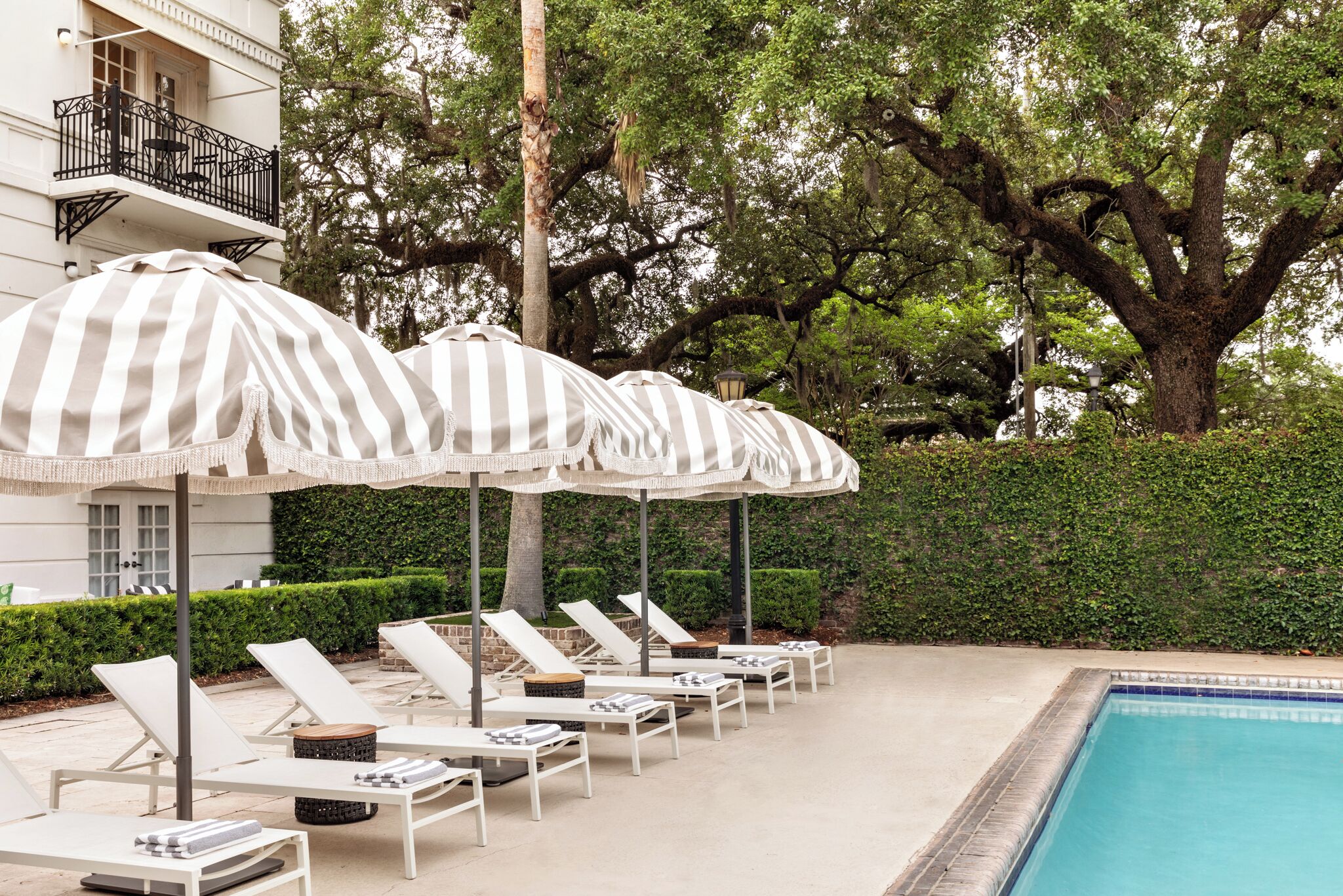 A poolside area with sun loungers and striped umbrellas, surrounded by trees and a hedge.
