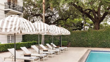A poolside area with sun loungers and striped umbrellas, surrounded by trees and a hedge.