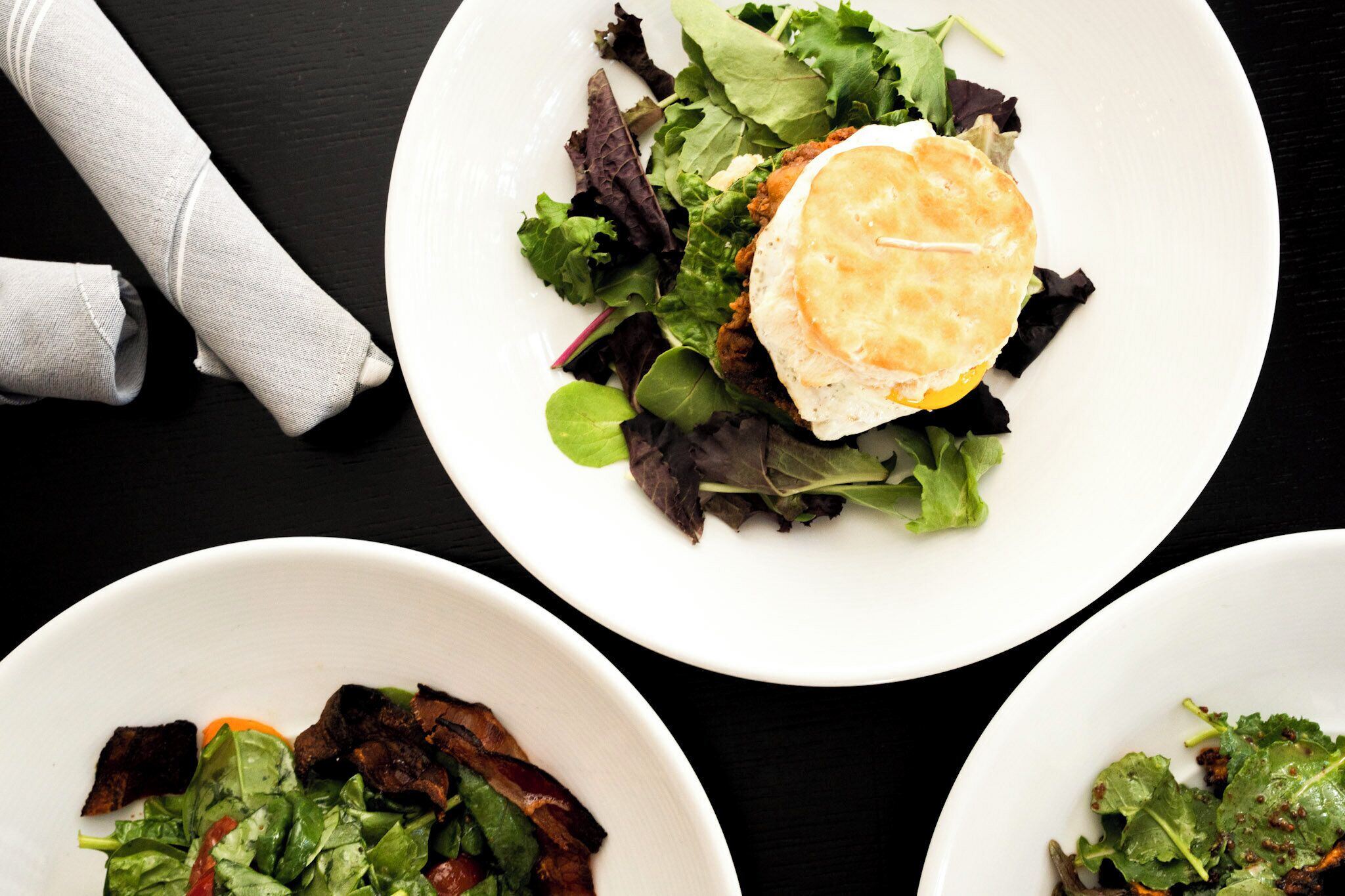 Tres platos blancos con ensaladas y una servilleta sobre una mesa negra.