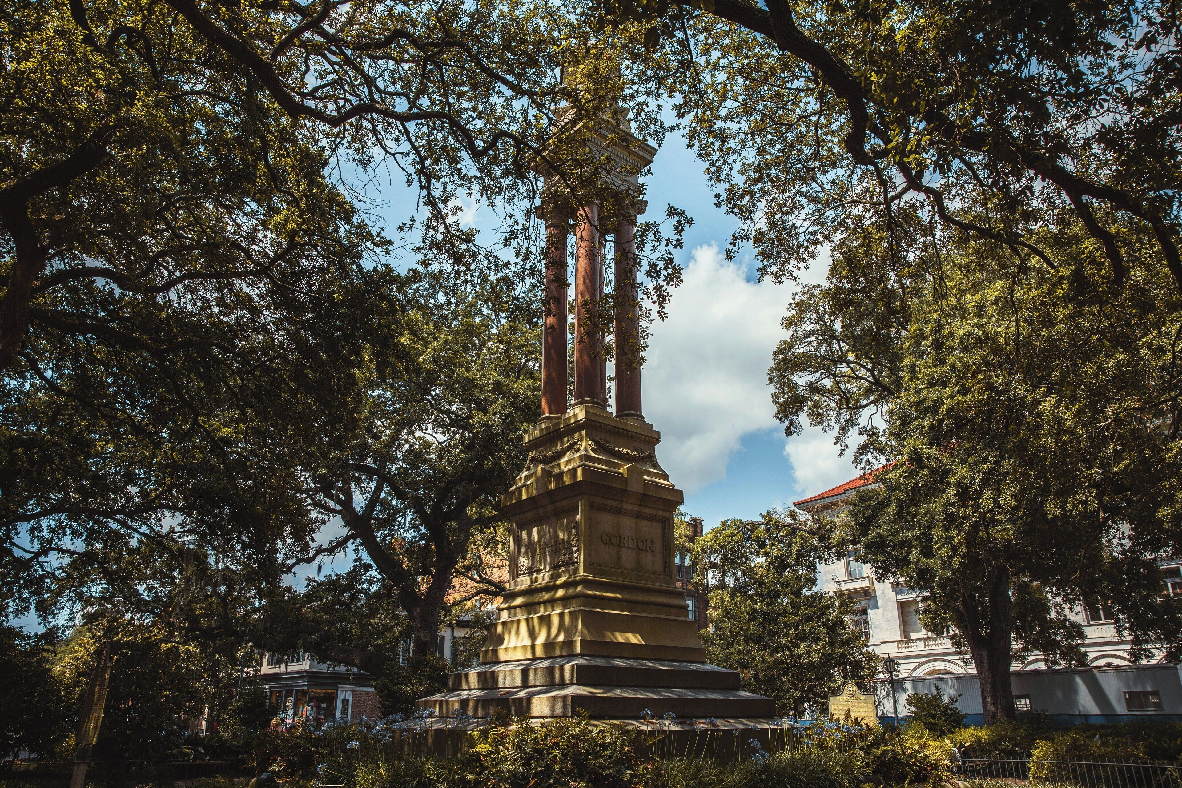 A historical monument with inscriptions, surrounded by lush trees and a building in the background.