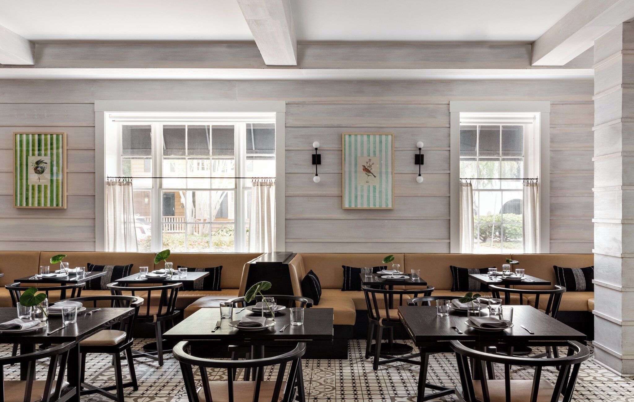 A dining area with multiple tables and chairs, a sofa, and two large windows with curtains.