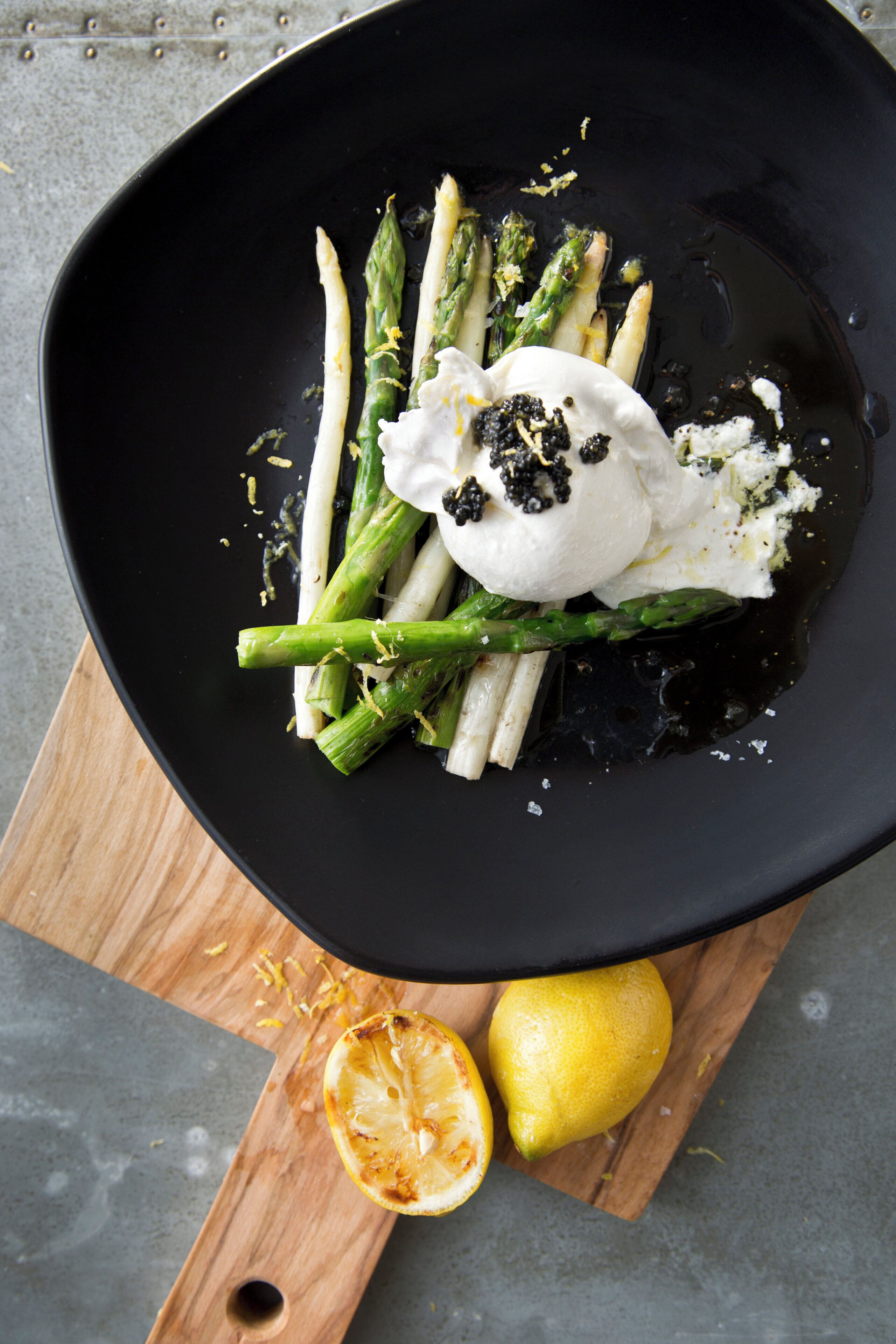 A black plate with roasted asparagus and a dollop of cream on a wooden board.