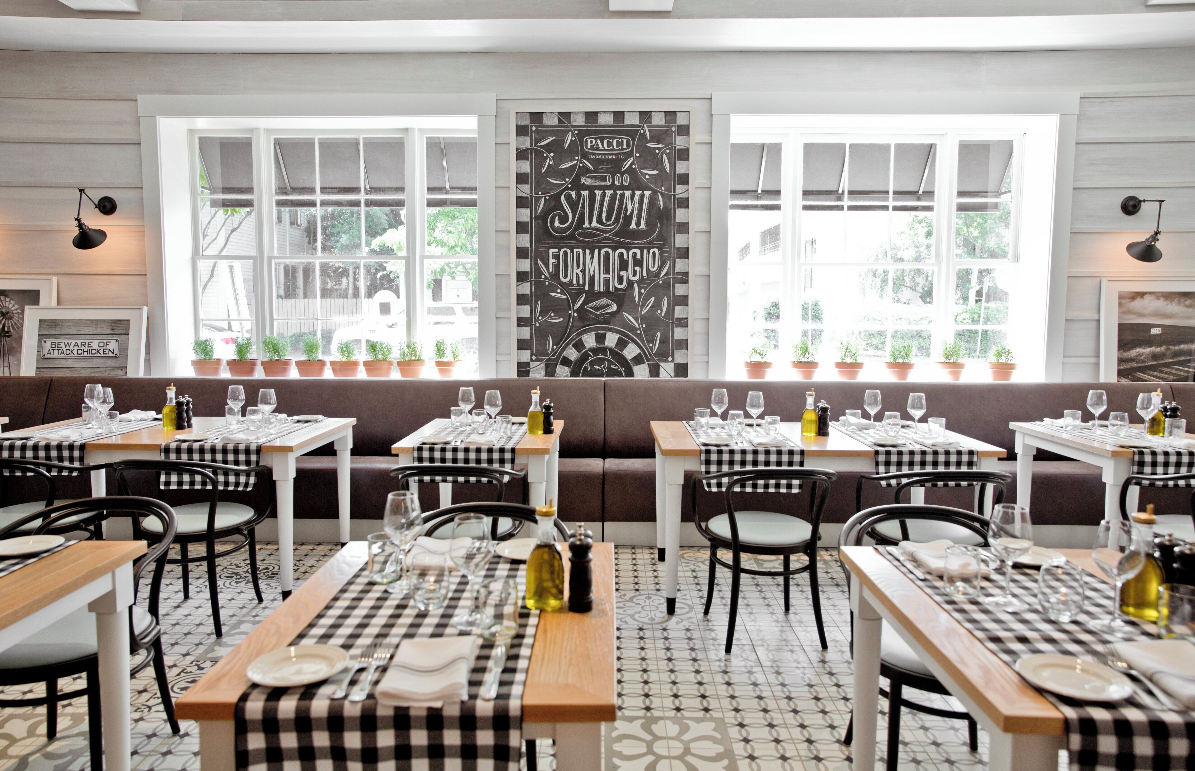 A restaurant interior with checkered tablecloths, black and white chairs, and a large window with potted plants.
